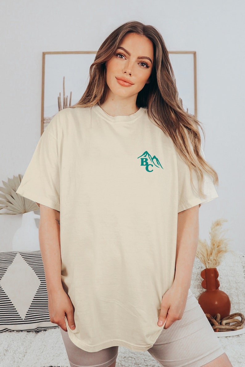 May include: Cream-colored t-shirt with a teal mountain graphic and the letters "BC". The shirt has short sleeves and a relaxed fit. The model is wearing neutral-colored shorts.