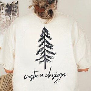 May include: Cream-colored t-shirt featuring a black graphic of a pine tree and the words "custom design" in a handwritten font. The shirt has short sleeves and a relaxed fit, suitable for casual wear.