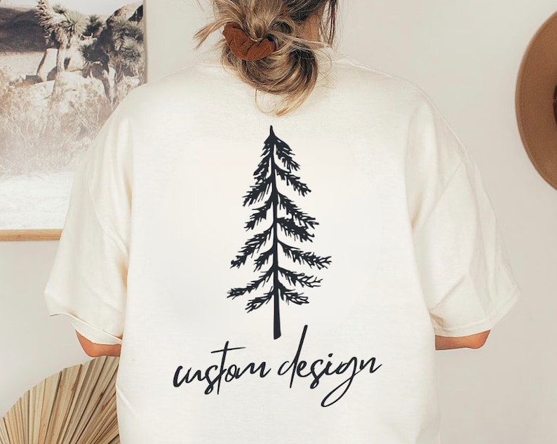 May include: Cream-colored t-shirt featuring a black graphic of a pine tree and the words "custom design" in a handwritten font. The shirt has short sleeves and a relaxed fit, suitable for casual wear.