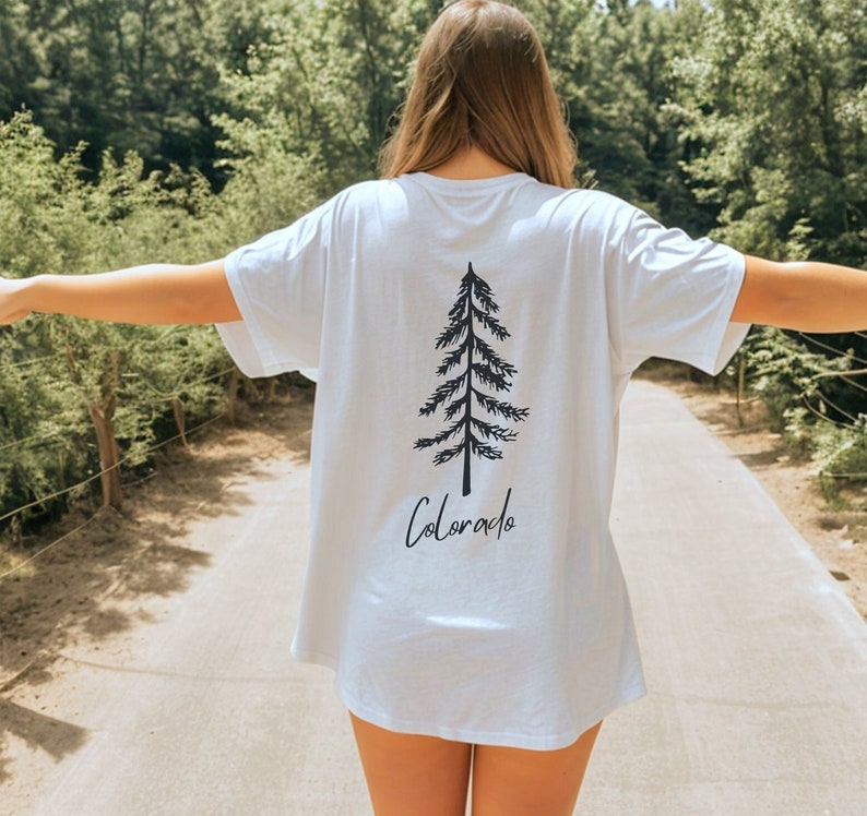 May include: White t-shirt featuring a black tree graphic and the word "Colorado" in a script font. The shirt has short sleeves and a relaxed fit. The image is taken outdoors with a blurred background of trees.
