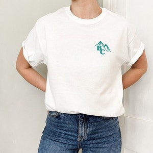 May include: White t-shirt with rolled-up sleeves and a teal mountain graphic with the letters "BC". The shirt is made of a soft material and is paired with blue jeans.