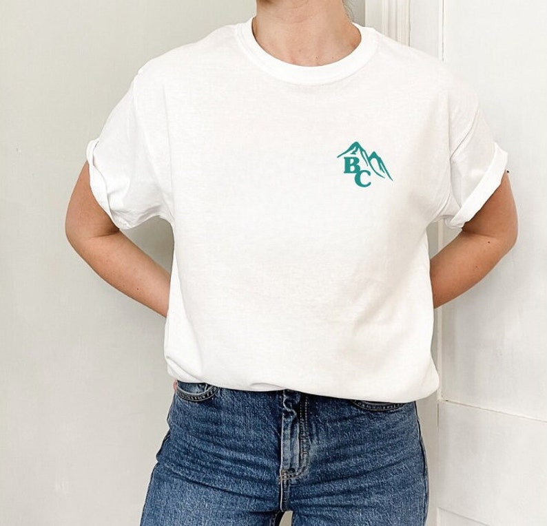 May include: White t-shirt with rolled-up sleeves and a teal mountain graphic with the letters "BC". The shirt is made of a soft material and is paired with blue jeans.