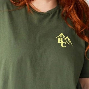 May include: Olive green t-shirt with short sleeves. The shirt features a yellow mountain graphic with the letters "BC" on the upper left chest. The shirt is made of a soft material and has a relaxed fit.