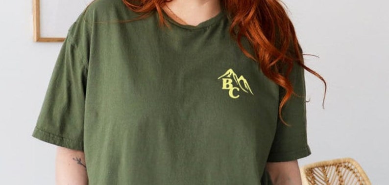 May include: Olive green t-shirt with short sleeves. The shirt features a yellow mountain graphic with the letters "BC" on the upper left chest. The shirt is made of a soft material and has a relaxed fit.
