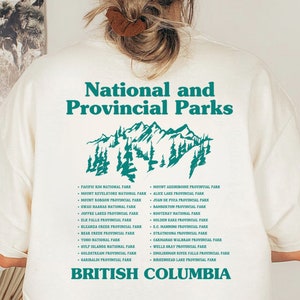 May include: Cream-colored t-shirt with a teal graphic of mountains and text. The text reads "National and Provincial Parks" above a list of park names and "British Columbia" below. A casual, nature-themed design.