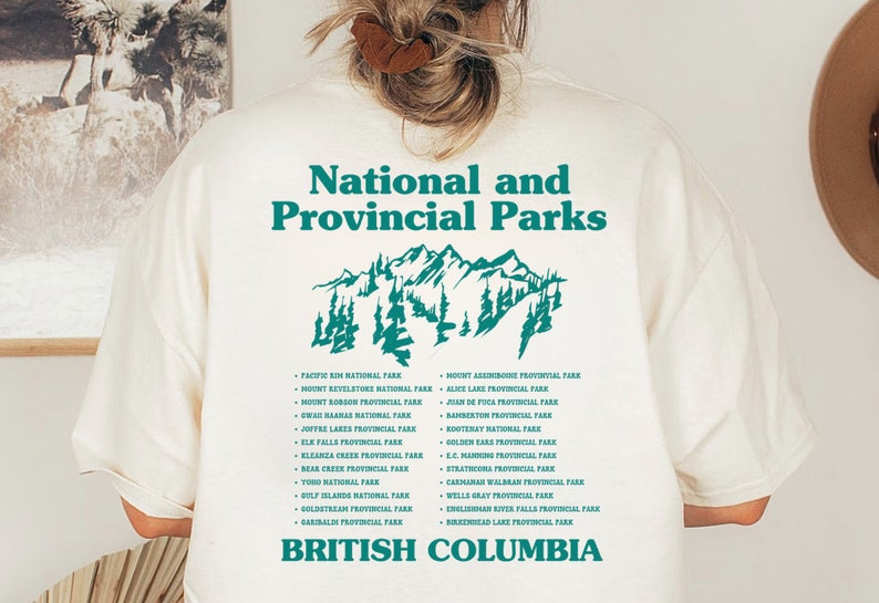 May include: Cream-colored t-shirt with a teal graphic of mountains and text. The text reads "National and Provincial Parks" above a list of park names and "British Columbia" below. A casual, nature-themed design.