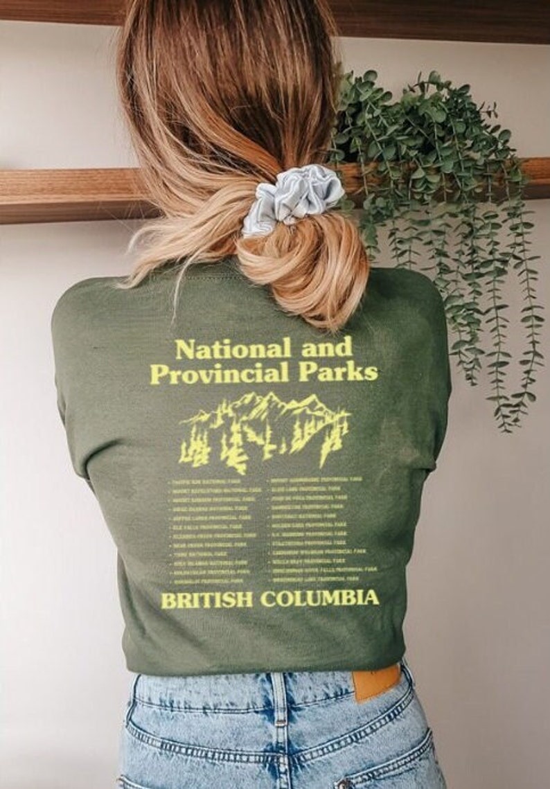 May include: Olive green t-shirt with the text "National and Provincial Parks" and "British Columbia" in yellow. A mountain graphic is centered between the text. The person is wearing blue jeans.