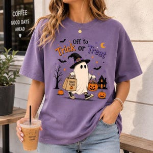 Könnte beinhalten: Ein violettes Kurzarm-T-Shirt mit einer Grafik eines Geistes, der einen Hexenhut und Turnschuhe trägt und eine Tasche mit der Aufschrift "Treats Please" hält. Das Design enthält den Text "Off to Trick or Treat" sowie Kürbisse, Fledermäuse und ein Haus.