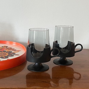 May include: Set of two vintage clear glass mugs with a smoky tint, each held in a black holder with a handle. The mugs sit on a wooden surface next to a floral patterned tray with an orange rim.