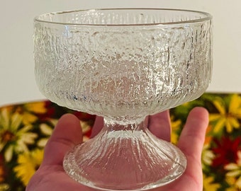 May include: A clear glass dessert bowl with a textured surface and a pedestal base. The bowl is held in a hand.