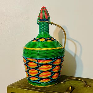 May include: A vintage, colorful woven pitcher with a handle. The pitcher is primarily green with accents of yellow, orange, red, and blue. The lid is cone-shaped and features red and green woven patterns. A gold-colored metal handle is attached.