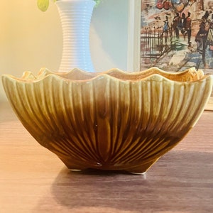 May include: A brown ceramic planter with a scalloped edge and a ribbed design. The planter is sitting on a wooden surface next to a vase of flowers and a framed picture.