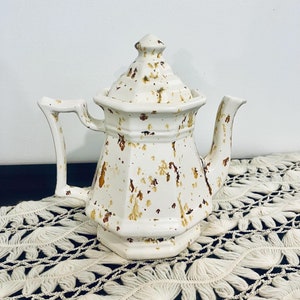May include: White ceramic teapot with a distressed, speckled brown glaze. The teapot has a rounded body and a lid with a knob on top. The teapot is sitting on a white crocheted doily.