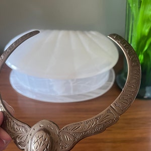 May include: A silver necklace with an ornate design. The necklace is made of a single piece of metal that is bent into a circle. The design features a central oval with a textured surface and intricate floral patterns on either side.