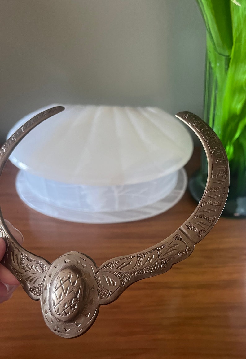 May include: A silver necklace with an ornate design. The necklace is made of a single piece of metal that is bent into a circle. The design features a central oval with a textured surface and intricate floral patterns on either side.