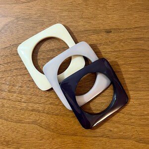 May include: Three square-shaped plastic bangles, one white, one gray, and one dark purple.