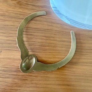 May include: A gold-toned metal bottle stopper with a decorative design. The stopper has a rounded top and a curved, open bottom.