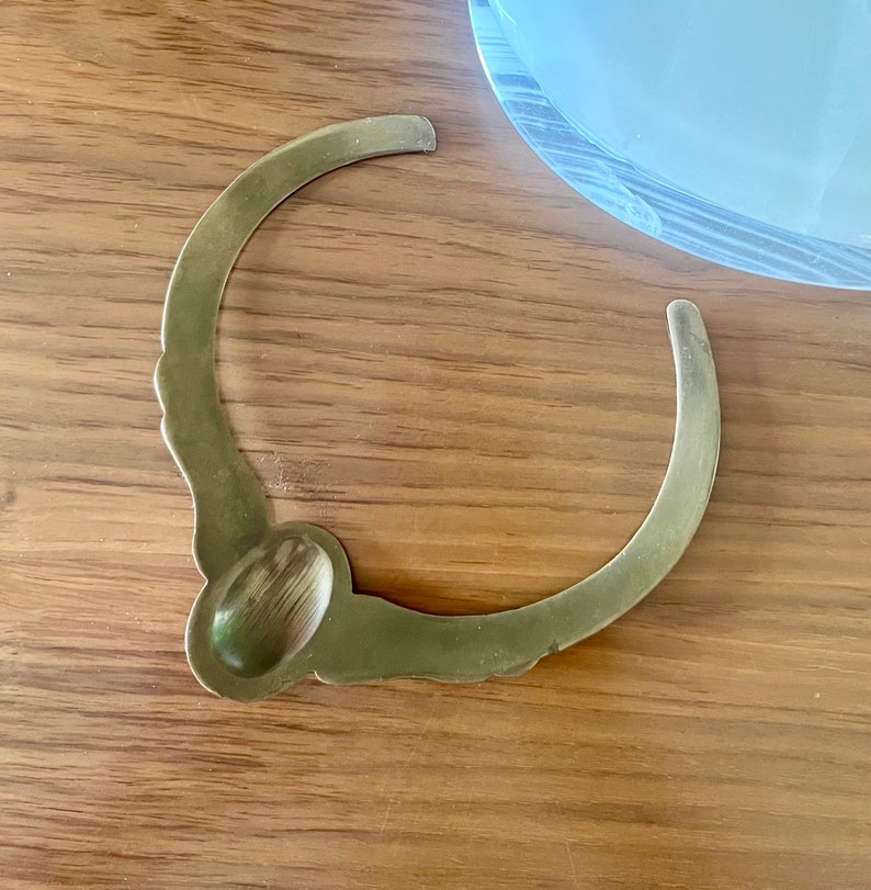 May include: A gold-toned metal bottle stopper with a decorative design. The stopper has a rounded top and a curved, open bottom.