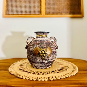May include: A decorative ceramic vase with two handles, featuring a textured, mottled brown and white finish. The vase is adorned with bronze-colored grape leaf and grape cluster embellishments. It sits on a round, beige woven placemat.