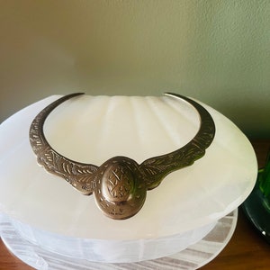 May include: A silver choker necklace with an ornate design. The necklace is made of metal and has a circular pendant with intricate details.