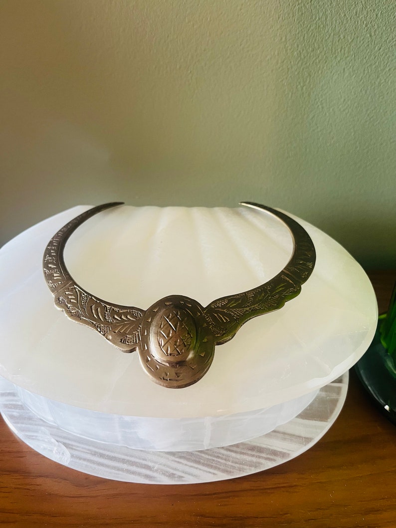 May include: A silver choker necklace with an ornate design. The necklace is made of metal and has a circular pendant with intricate details.