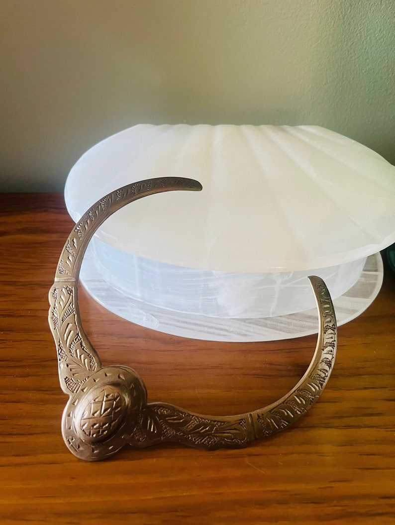 May include: A silver-toned metal bracelet with an intricate design. The bracelet is curved and has a decorative clasp.