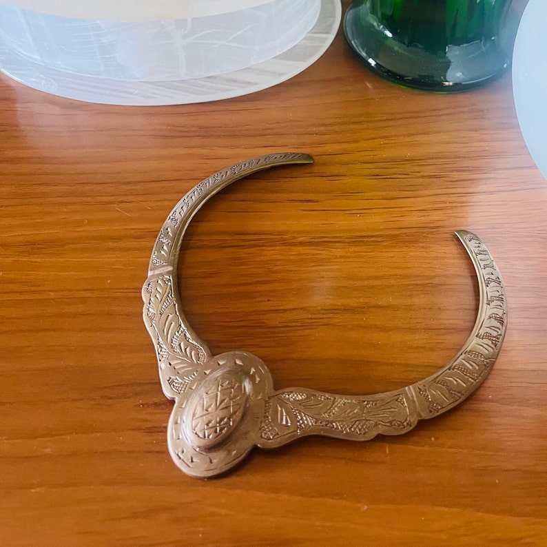 May include: A silver-toned metal necklace with an ornate design. The necklace is shaped like a crescent moon and has a large, oval-shaped pendant in the center.