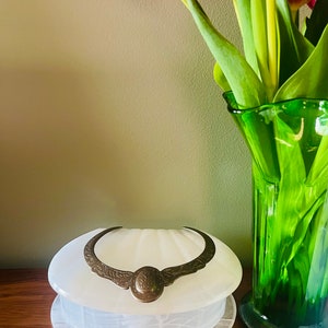 May include: A white alabaster shell-shaped trinket box with a gold-toned metal hinge and a decorative design. The box is sitting on a wooden surface next to a green glass vase with pink tulips.