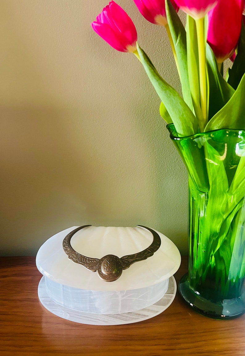 May include: A white alabaster shell-shaped trinket box with a gold-toned metal hinge and a decorative design. The box is sitting on a wooden surface next to a green glass vase with pink tulips.