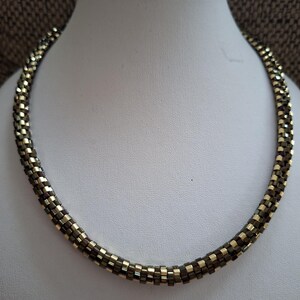 May include: A beaded necklace displayed on a white mannequin. The necklace is a tube shape, with alternating gold and dark brown beads. The beads are small and square, creating a textured surface. The necklace is curved, suggesting it is meant to be worn around the neck.