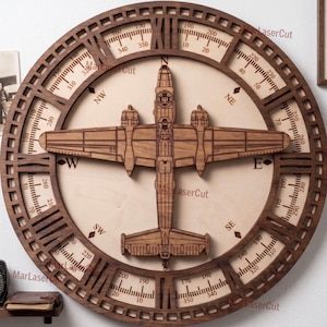 Puede incluir: Decoración de pared circular de madera con un diseño detallado de avión en el centro, rodeado por una rosa de los vientos. El anillo exterior muestra marcas de grados e indicadores direccionales. La pieza es de madera marrón clara y oscura.
