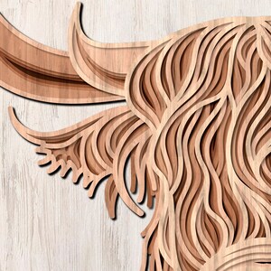 May include: A close-up of a wooden cutout of a Highland cow's head. The cow's fur is depicted with intricate layered lines, creating a 3D effect. The wood is a light brown color.