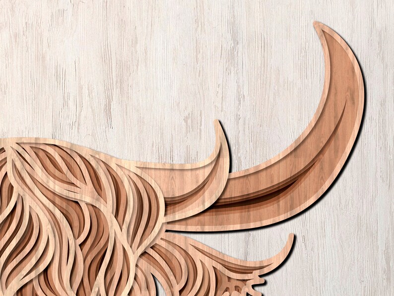 May include: A wooden cutout of a Highland cow with intricate layered details. The cow's head is facing the left side of the image, and the horns are curled up and out. The background is a light wood grain.