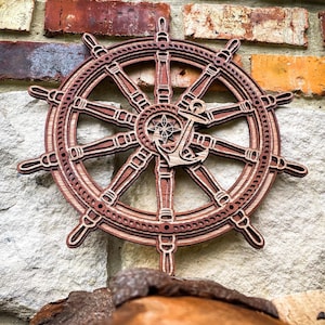 May include: A wooden ship's wheel wall decoration with a nautical theme. The wheel is made of layered wood in shades of brown and tan, featuring an anchor and compass rose design in the centre. The wall decoration is mounted on a stone and brick wall.