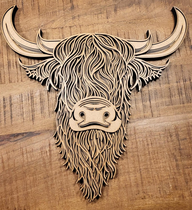 May include: A wooden cutout of a Highland cow head with intricate details in the fur and horns. The cow's face is detailed with a black nose and eyes.