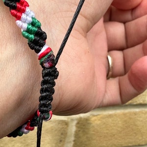 May include: A black and green friendship bracelet with red, white, and green beads. The bracelet is tied around a person's wrist.