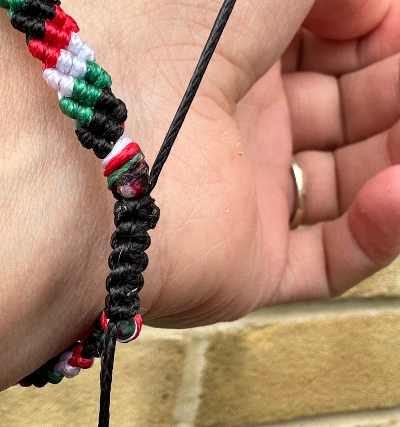 May include: A black and green friendship bracelet with red, white, and green beads. The bracelet is tied around a person's wrist.