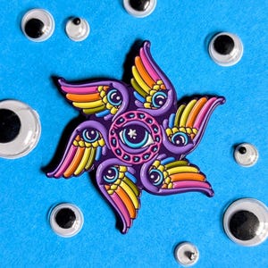 May include: A colorful enamel pin featuring a starburst design with multiple eyes and wings. The pin is surrounded by googly eyes on a blue background.