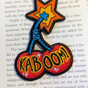 Puede incluir: Un parche bordado rojo y amarillo con dos cerezas y la palabra "KABOOM" en letras negras. Las cerezas tienen estrellas blancas.