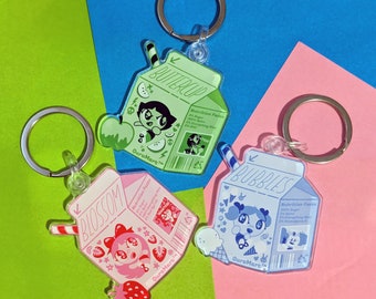 May include: Three cartoon character themed keychains shaped like milk cartons. The keychains are pink, blue, and green with the words "Blossom", "Bubbles", and "Buttercup" on them. Each keychain has a cartoon character on the front and nutritional information on the back.