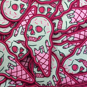 Puede incluir: Un montón de parches bordados de color rosa y verde que representan una calavera derritiéndose en un cono de helado. El cono de helado está decorado con un patrón de cono de gofre rosa y blanco y con chispas rosas.