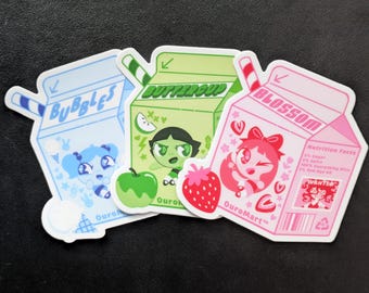May include: Three die-cut stickers shaped like milk cartons in light blue, light green, and light pink. Each features a Powerpuff Girl character, a straw, fruit graphics, and text including Bubbles, Buttercup, Blossom, and OuroMart.