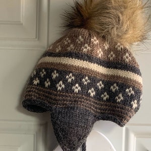May include: A brown and white knitted hat with a brown faux fur pom pom. The hat has a fold-down ear flap and a tie string.