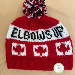 Peut inclure: Toque rouge en tricot avec un pompon blanc et des rayures noires et blanches. La toque a une bande blanche avec les mots "Elbows Up" en lettres noires. Il y a trois motifs de feuilles d'érable canadiennes blanches sur la toque. La toque a une petite étiquette verte avec les mots "Hush I'm Creating" en lettres noires.