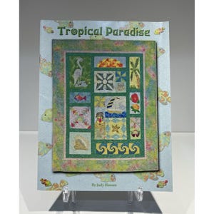 May include: A pattern book titled Tropical Paradise by Judy Hansen featuring a quilt design with various tropical motifs including a heron, sailboat, palm tree, flowers, and seashells, set against a light blue background with fish illustrations.