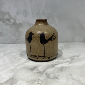 May include: A small, round, tan ceramic vase with a speckled texture. The surface features a black graphic illustration of two birds facing each other, with thin legs and long tails. The vase has a narrow neck and a wide, rounded body.