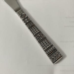 May include: A stainless steel knife handle featuring a series of engraved geometric patterns including diamonds, four-petal flowers, chevrons, triangles, and infinity loops arranged in horizontal bands along the flat, rectangular surface.