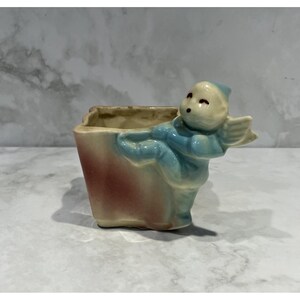 May include: A ceramic planter featuring a sculpted figure of a small winged being in a light blue finish. The main body of the planter has a cream-colored base with a soft, blended reddish-brown gradient on the side, creating a distinct contrast.