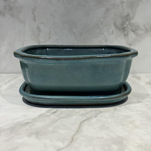 May include: A teal ceramic rectangular planter with a matching saucer. The planter has a slightly flared shape with a dark brown rim. The saucer is a simple, rectangular shape. The set is glazed with a glossy finish.