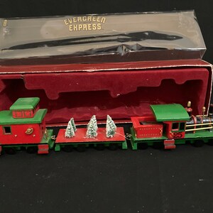 May include: A wooden toy train set with a red caboose, a flatbed car carrying three small artificial Christmas trees, and a green engine with the number 25 on the side. The train is in its original box with the words "Evergreen Express" printed on the top.